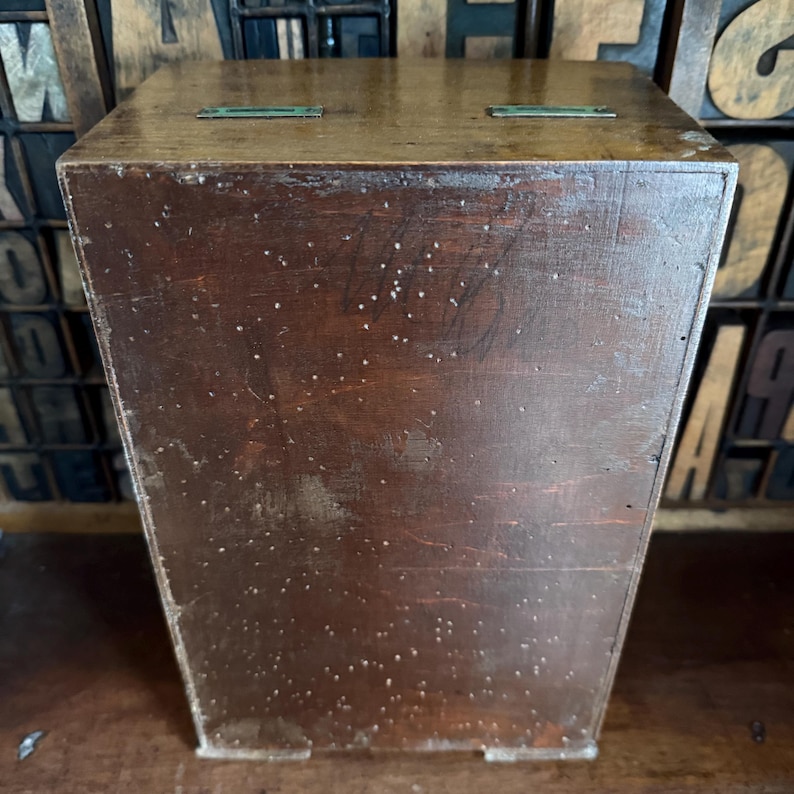 May include: A vintage wooden box with a dark brown finish. The box has a rectangular shape with a slightly tapered design. Two small, rectangular metal accents are visible on the top. The wood shows signs of wear, adding to its antique aesthetic.