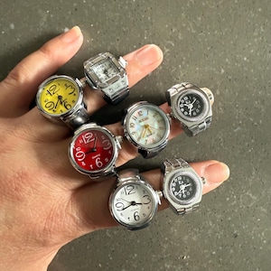May include: A collection of ring watches with various colored faces, including yellow, red, white, and black. The rings have silver-toned bands and feature quartz movement. The watch faces display numbers and hands for telling time.
