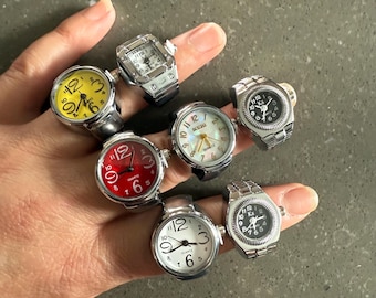 May include: A collection of ring watches with various colored faces, including yellow, red, white, and black. The rings have silver-toned bands and feature quartz movement. The watch faces display numbers and hands for telling time.