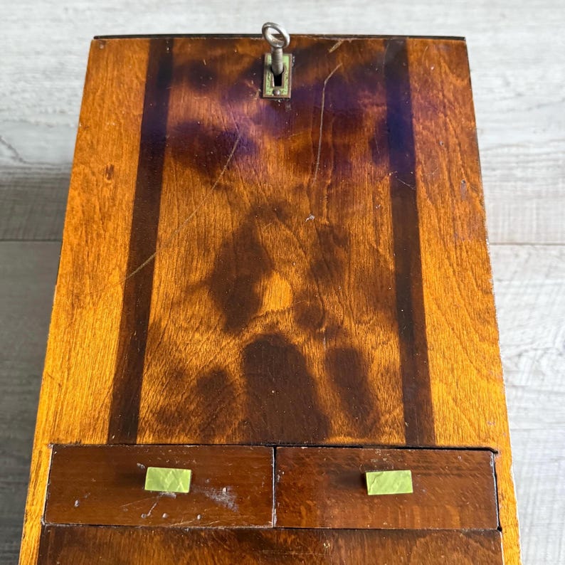 May include: A vintage wooden desk with a keyhole and two small drawers. The desk is made of dark and light brown wood with a glossy finish. The drawer pulls are a light green color.