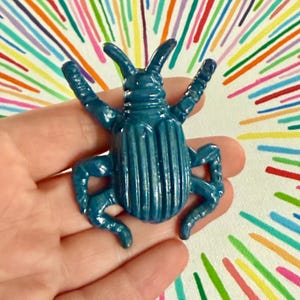 May include: A teal-colored, stylized beetle figurine with detailed ridges on its back and legs. The beetle is held in a hand against a background of colorful, radiating lines.