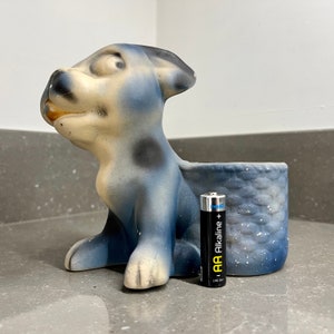 Könnte beinhalten: Eine blaue und weiße Keramik-Hundefigur mit schwarzen Flecken. Der Hund sitzt mit dem Kopf zur Seite geneigt und leicht geöffnetem Maul. Die Figur hat einen kleinen Korb auf dem Rücken, der ebenfalls blau und weiß mit einem gewebten Muster ist. Eine schwarze AA-Batterie liegt im Korb.