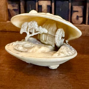 Könnte beinhalten: Eine detaillierte elfenbeinfarbene Skulptur einer Miniaturlandschaft in einer offenen Muschel. Die Szene umfasst ein kleines Haus, einen Baum und ein Wasserrad. Die Skulptur steht auf einer Holzoberfläche.