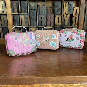 May include: Three miniature suitcases with different designs. The first suitcase is pink with a brown leather texture and has various travel stickers. The second suitcase is brown with a map design and has a "TRIP" label. The third suitcase is pink with a floral design and a bird with the text "Perfect Quality".