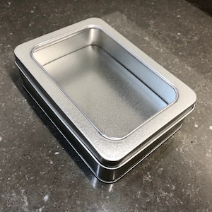 May include: A rectangular, silver-colored tin box with a clear window in the lid. The box has a shiny, metallic finish and a simple, clean design. The window allows a view of the contents inside.