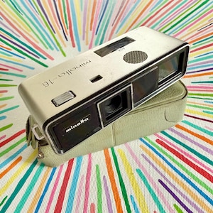 May include: A silver Minolta-16 subminiature camera with a black lens housing and a rectangular viewfinder. The camera features a silver metal body with the text Minolta-16 and Minolta printed on the front, resting on a light green case.