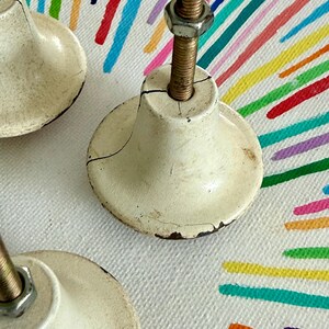 May include: Three off-white, bell-shaped cabinet knobs with a distressed, cracked finish. Each knob features a central threaded metal bolt extending upwards, topped with a hexagonal metal nut. The knobs have a wide, circular base and a tapered neck.