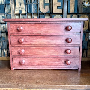 May include: A small, four-drawer wooden chest with a reddish-brown finish. Each drawer has a round, wooden knob. The chest is made of wood and has a simple, classic design. The chest is suitable for storing small items.