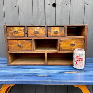 以下が含まれることがあります： 複数の引き出しと棚が付いたヴィンテージの木製キャビネット。キャビネットは薄い茶色の木でできており、素朴な外観をしています。青い木製の表面の上に置かれています。