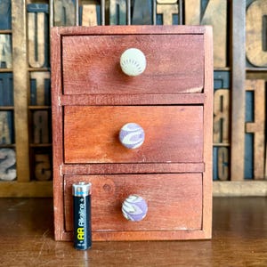 May include: A small, three-drawer wooden chest with a reddish-brown finish. Each drawer has a fabric-covered knob. An AA battery is placed in front of the chest for size comparison.
