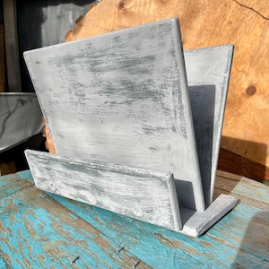 May include: A white wooden cookbook stand with a distressed finish. The stand is made of two pieces of wood that are joined together at a right angle. The stand is sitting on a blue wooden surface.
