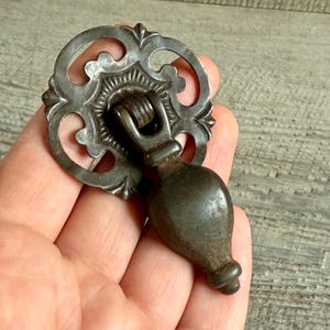 Könnte beinhalten: Antiker Metall-Schubladengriff mit einer verzierten, kreisförmigen Rückplatte und einem tropfenförmigen Griff. Der Griff hat eine dunkle, gealterte Oberfläche, die aufwendige Schnörkel und eine Vintage-Ästhetik zeigt.