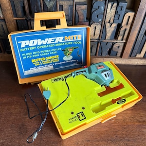 May include: An orange Power Mite miniature tool set with a green buffer-sander. The set includes a carry case with the text "Power Mite" and "Battery Operated Miniature Tool". The set includes a buffer-sander with 1 polish bonnet and 3 sanding discs.