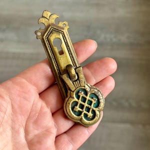 May include: Vintage brass door pull with a keyhole and decorative floral detailing. The pull has a rectangular backplate with a keyhole and a decorative handle. The handle features a green enamel inlay and a quatrefoil design.
