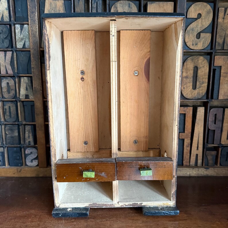 May include: A small, vintage wooden cabinet with two drawers and two compartments. The cabinet is made of light-colored wood with a dark-colored top and base. The drawers have green handles. The cabinet is likely used for storage.