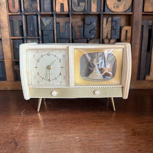 May include: Vintage ivory-colored clock radio with a clock face on the left and a miniature scene on the right. The clock has a gold-tone bezel and legs. The clock face has black numerals and hands. The miniature scene depicts a couple dancing.