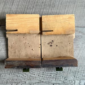May include: Two wooden blocks with a light brown top, a medium brown middle section, and a dark brown base. The blocks are connected and rest on green metal supports. The wood has visible grain and some small holes.