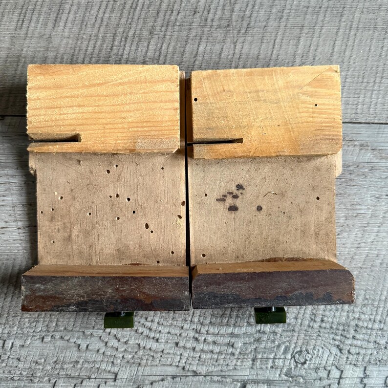May include: Two wooden blocks with a light brown top, a medium brown middle section, and a dark brown base. The blocks are connected and rest on green metal supports. The wood has visible grain and some small holes.