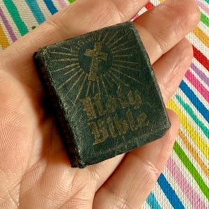 May include: A miniature dark green book with a textured cover featuring a gold-colored cross and sunburst design above the text Holy Bible. The small rectangular volume has visible wear along the edges and spine, showcasing a compact, handheld size.