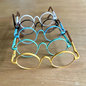 May include: Five pairs of round-framed spectacles in a stack, with frames in white, black, light blue, teal, and yellow. The temples are brown or yellow. The lenses are clear.