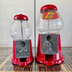 May include: Two red candy dispensers with clear glass globes and silver-toned metal bases featuring leaf patterns. The larger dispenser displays the Jelly Belly logo and the text The original gourmet jelly bean on the front of the glass globe.