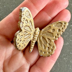 May include: A gold-colored butterfly-shaped decorative object. The butterfly has detailed wing patterns and a textured surface. The object is held in a hand, showcasing its size and design.