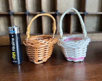 May include: Two small woven baskets with handles. The basket on the left is brown and the basket on the right is white with a pink trim.