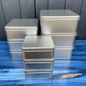 May include: A stack of four silver metal rectangular tins with lids. The tins are of varying sizes, with the largest at the top and the smallest at the bottom. The tins are arranged on a blue wooden surface.