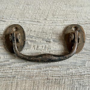 Könnte beinhalten: Ein antiker Metall-Schubladengriff mit ovalen Grundplatten und einem gebogenen Griff. Die Beschläge weisen Alterserscheinungen wie Rost und Abnutzung auf, was ihnen ein antikes Aussehen verleiht.