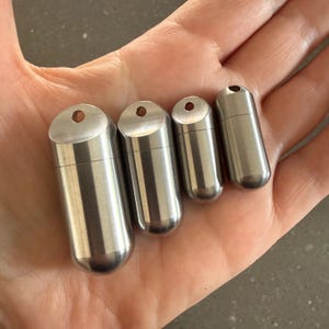 May include: Four cylindrical metal capsules of varying sizes, each featuring a rounded bottom and a flat top with a small circular hole for attachment. The capsules have a smooth, brushed silver finish and a horizontal seam indicating a screw-top lid.