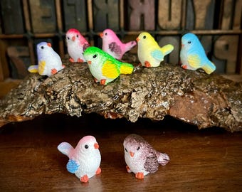 May include: A collection of eight small, colorful bird figurines. The birds are made of ceramic and are painted in various shades of pink, blue, yellow, green, and white. The birds are arranged on a piece of tree bark.