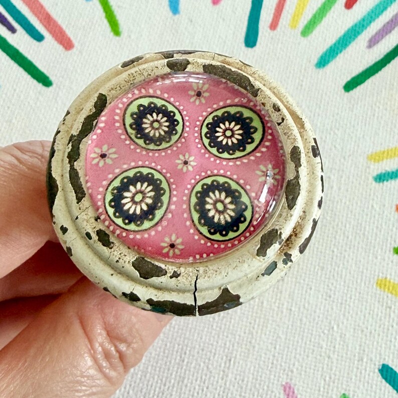 May include: A round cabinet knob featuring a distressed white painted metal frame with visible dark underlayers. The center is a circular glass inlay displaying a pink background patterned with four green and dark blue floral motifs.
