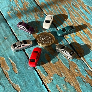 May include: A collection of miniature toy cars in various colors, including silver, red, purple, and blue, arranged in a circle on a blue and brown wooden surface. A British one pound coin is in the center of the circle.