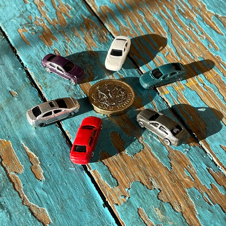 May include: A collection of miniature toy cars in various colors, including silver, red, purple, and blue, arranged in a circle on a blue and brown wooden surface. A British one pound coin is in the center of the circle.