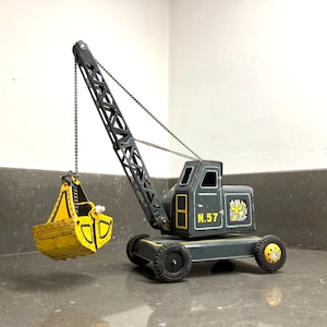 May include: A vintage toy crane with a yellow bucket, black chains, and a gray metal body. The crane has a yellow and black number plate that reads "N.57".