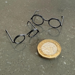 May include: Two pairs of small, round, black wire-frame glasses sit next to a one-pound coin. The glasses have circular lenses and straight temples. The coin is gold and silver in color.