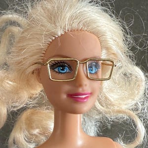 May include: A blonde doll wearing gold-rimmed square glasses with blue lenses.