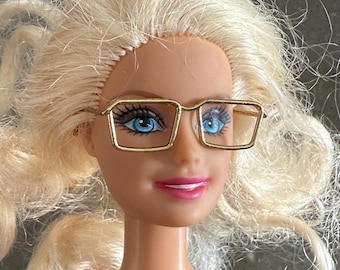 May include: A blonde doll wearing gold-rimmed square glasses with blue lenses.