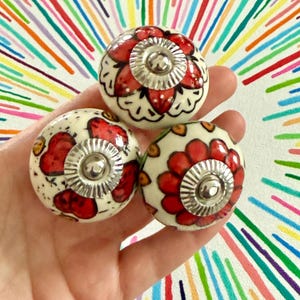 May include: Three ceramic cabinet knobs with a floral design. The knobs are off-white with red flower details and silver metal accents. The knobs are held in a hand against a colourful background.