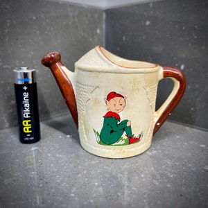 May include: A small, white ceramic watering can with a brown handle and spout. The can features a hand-painted illustration of a green elf sitting on a bed of green leaves. The elf is wearing a red hat and red shoes.