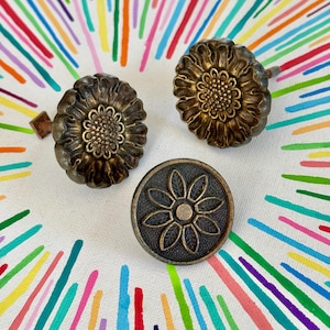 May include: Two antique brass-toned drawer knobs shaped like blooming sunflowers with textured centres and one flat, circular brass-toned knob featuring a raised, eight-petaled flower design with a stippled background texture.