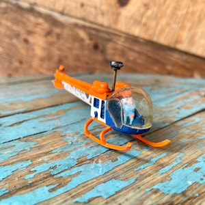 Könnte beinhalten: Ein Spielzeughelikopter mit orangefarbenen und blauen Details. Der Helikopter hat einen durchsichtigen Plastik-Cockpit mit einer kleinen Figur darin. Der Helikopter steht auf einer Holzoberfläche.