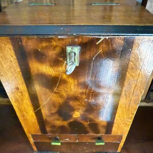 May include: A vintage wooden chest with a glossy finish. The chest features a dark brown top and a lighter brown body with vertical wood grain. It has a metal latch and two small drawers at the bottom with green accents.