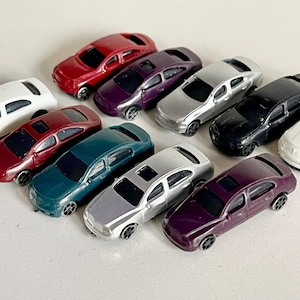Könnte beinhalten: Zehn Miniatur-Spielzeugautos in verschiedenen Farben, darunter Rot, Lila, Silber, Schwarz, Weiß und Grün. Die Autos sind alle gleich groß und haben die gleiche Form, und sie sind in einem Halbkreis auf einer weißen Oberfläche angeordnet.