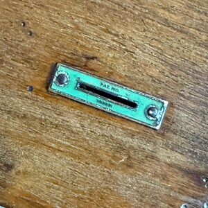 May include: Close-up of a rectangular, green metal slot with the text "PAT. NO." and other illegible text. The slot is set into a wooden surface, with two rivets on either end.
