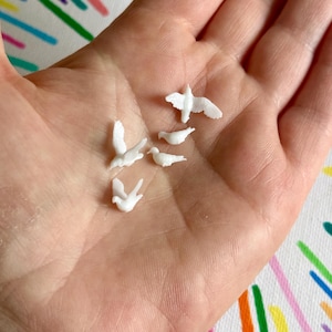 May include: A selection of tiny, white dove ornaments in different poses, including flying and resting. The miniature birds are held in an open hand, highlighting their intricate details and forms. The background features a colourful, patterned fabric.
