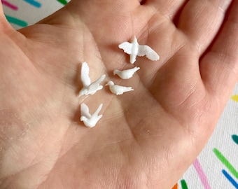 May include: A collection of small, white dove figurines in various poses, including flying and perched positions. The miniature birds are displayed in an open hand, showcasing their delicate details and shapes. The background is a colorful, patterned fabric.