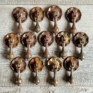 May include: A selection of antique bronze drawer pulls. Each pull has a circular backplate with a decorative edge and a teardrop-shaped handle. The pulls have a weathered, aged look, with colour variations from bronze to gold.