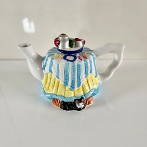 Könnte beinhalten: Eine weiße Teekanne aus Keramik mit einem Design aus blau-gelb gestreiftem Tischtuch. Die Teekanne hat eine schwarze Katzenfigur am Boden und eine kleine Teetasse und Teekanne oben drauf.
