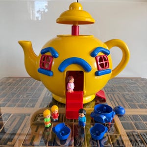 May include: A yellow plastic teapot-shaped playhouse with a red door, blue window frames, and a removable lid. The set includes three small plastic figures and two blue toy cups, with one figure standing on the red stairs of the teapot house.